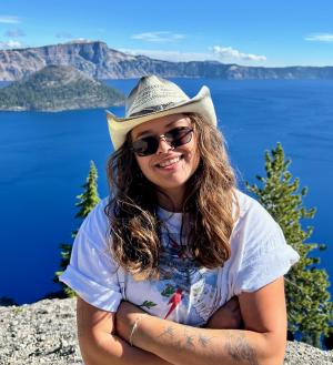 Audrey, a brown haired woman, stands with arms crossed in front of a lake wearing sunglasses and a cowboy hat.