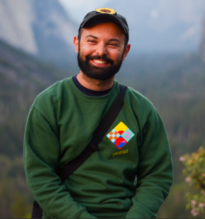 Abdurrafey, a dark haired man with a beard, smiles at the camera in front of a green background